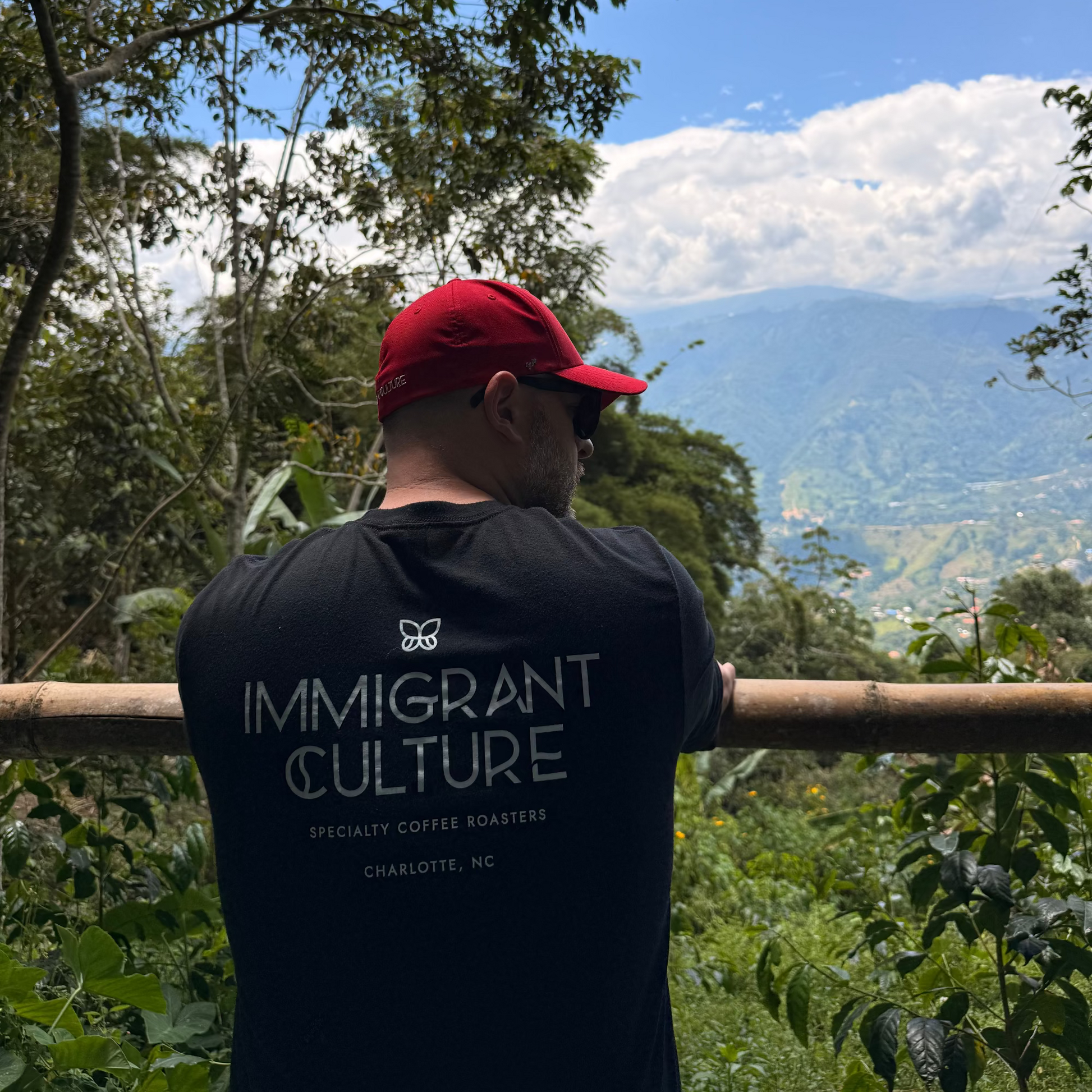 Person wearing a 'Immigrant Culture' t-shirt with a scenic view of mountains and trees.