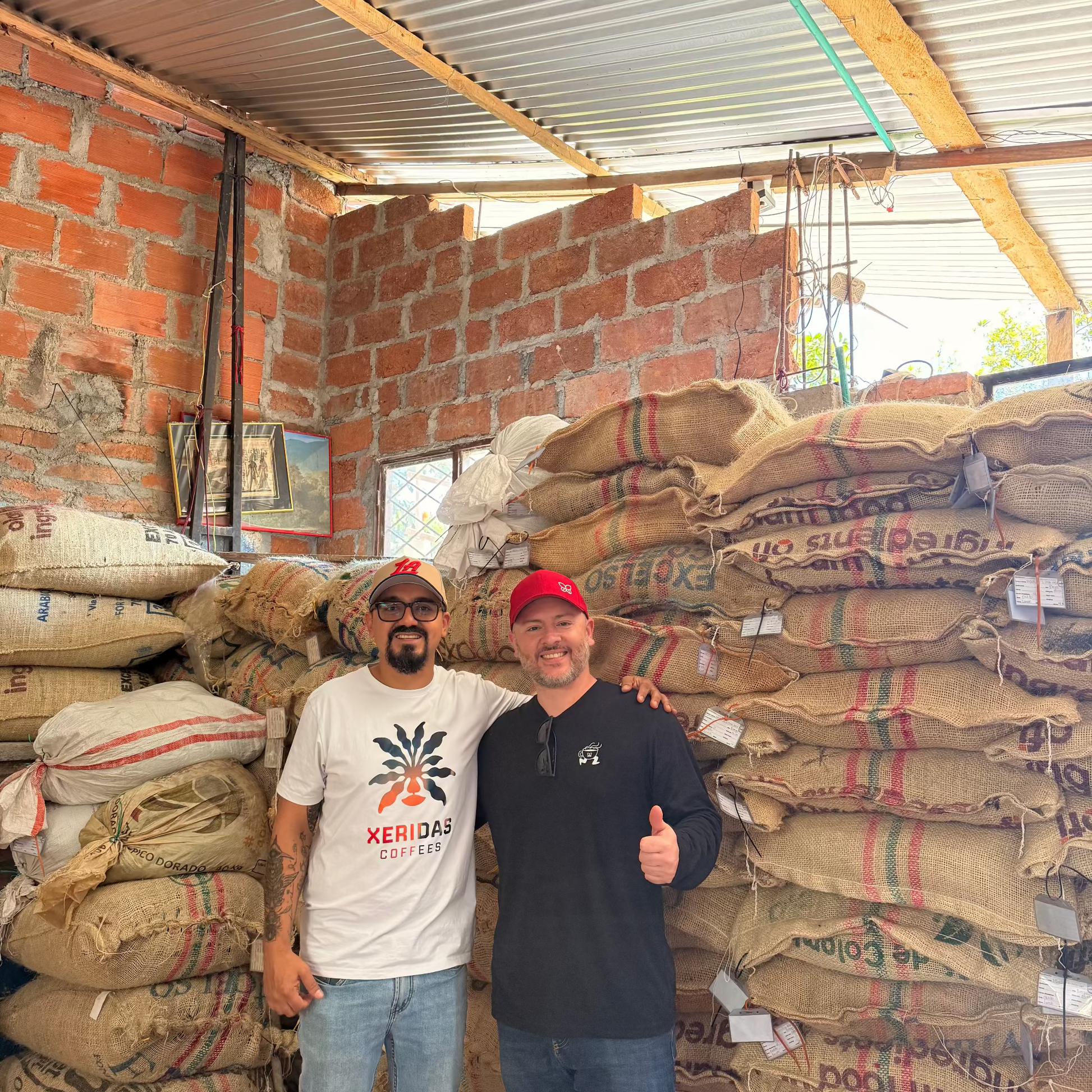 Gonzalo and Maurizio standing in a warehouse at San Fernando Coffee farm with stacked bags of coffee.