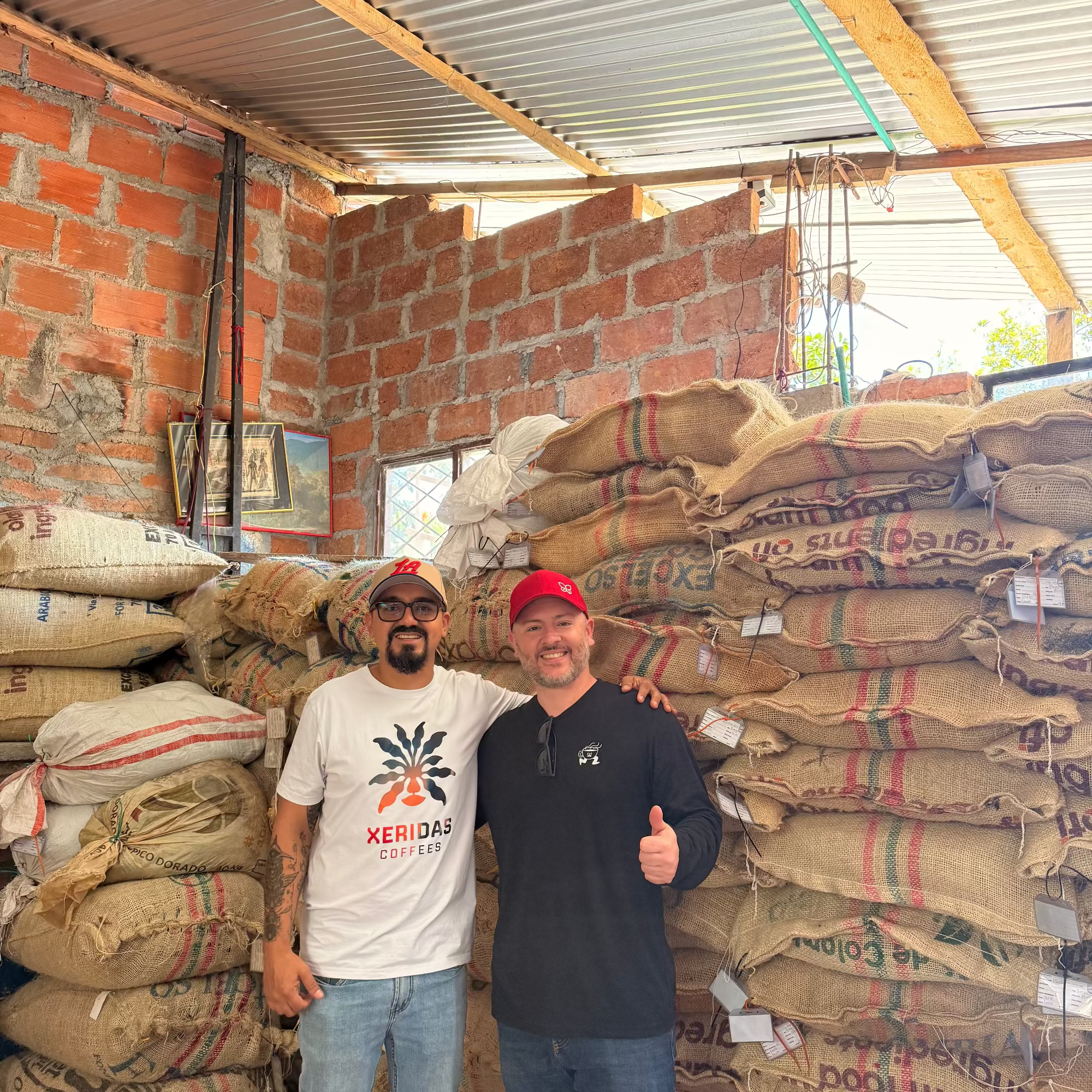 Gonzalo and Maurizio standing in a warehouse at San Fernando Coffee farm with stacked bags of coffee.